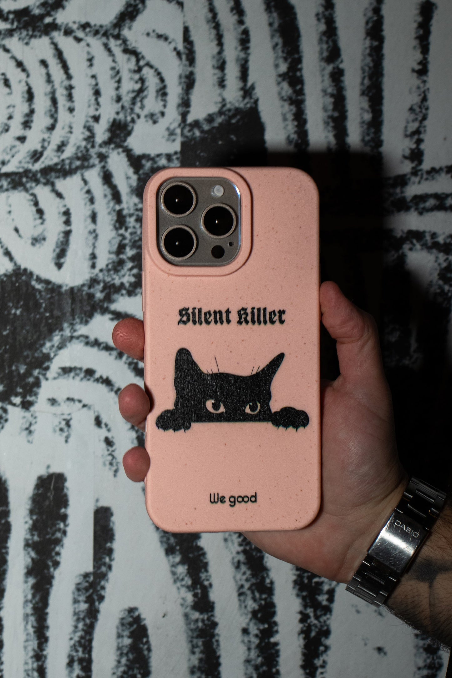 Man holding a We Good Tpu iPhone case with wheat mix speckled in a flamingo pink color with a black 'silent killer' text in a gothic font and below it a black cats face creeping up and stalking.