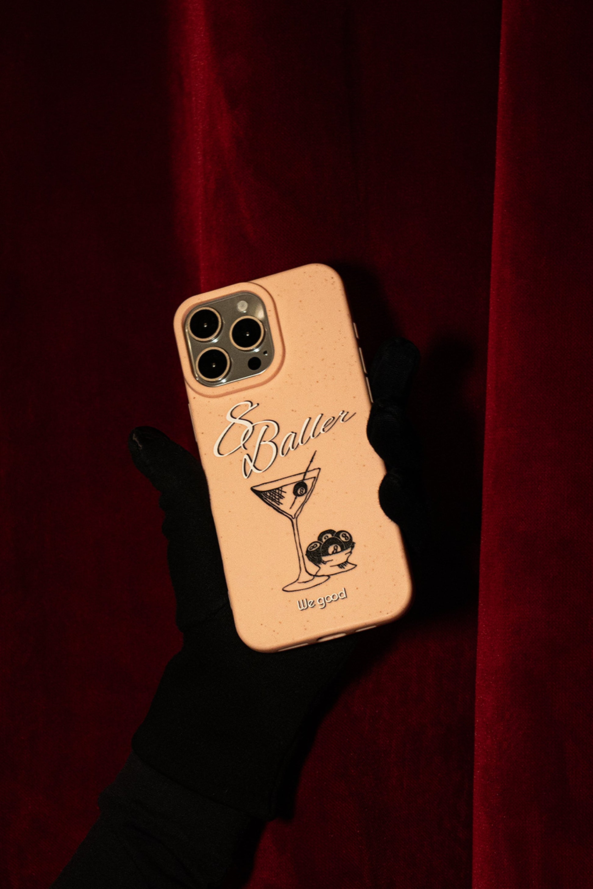 Woman with black gloves holding a We Good Tpu iPhone case with wheat mix speckled in a flamingo pink color with '8 baller' white script text print and a black print martini glass with pool 8 balls. the phone is being held in front of a red velvet curtain.