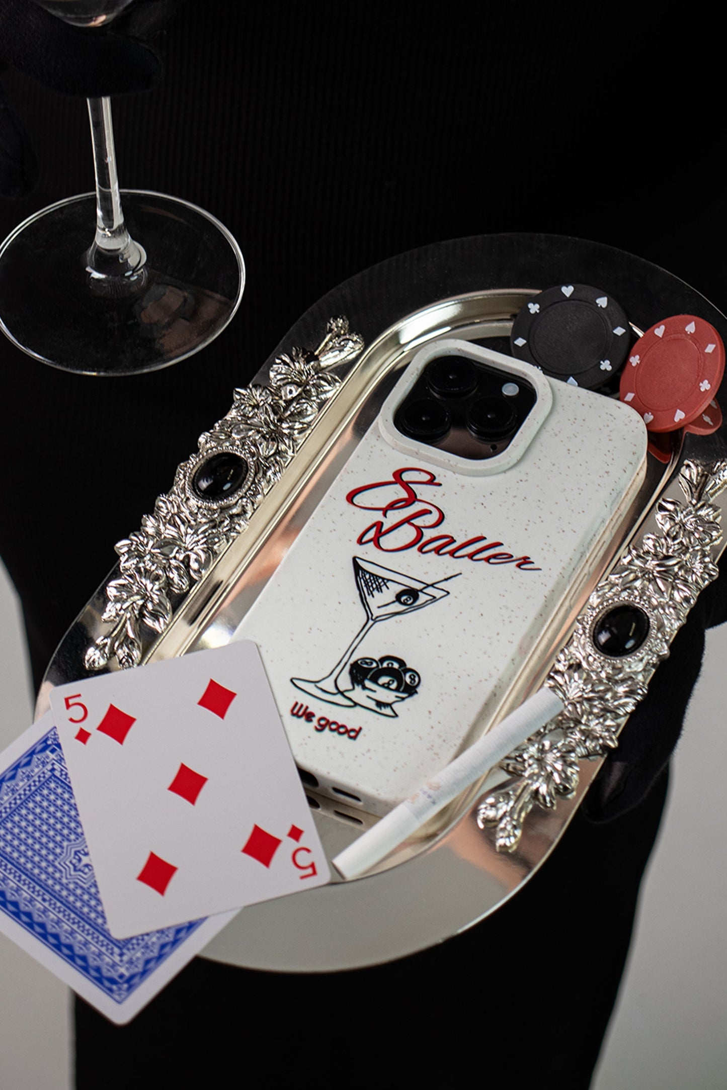 woman in a black dress holding a martini glass and silver tray with playing cards, a cigarette and a We Good Tpu iPhone case with wheat mix speckled in an off-white color with '8 baller' red script text print and a black print martini glass with pool 8 balls.