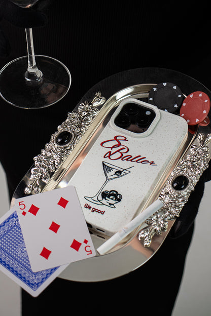 woman in a black dress holding a martini glass and silver tray with playing cards, a cigarette and a We Good Tpu iPhone case with wheat mix speckled in an off-white color with '8 baller' red script text print and a black print martini glass with pool 8 balls.