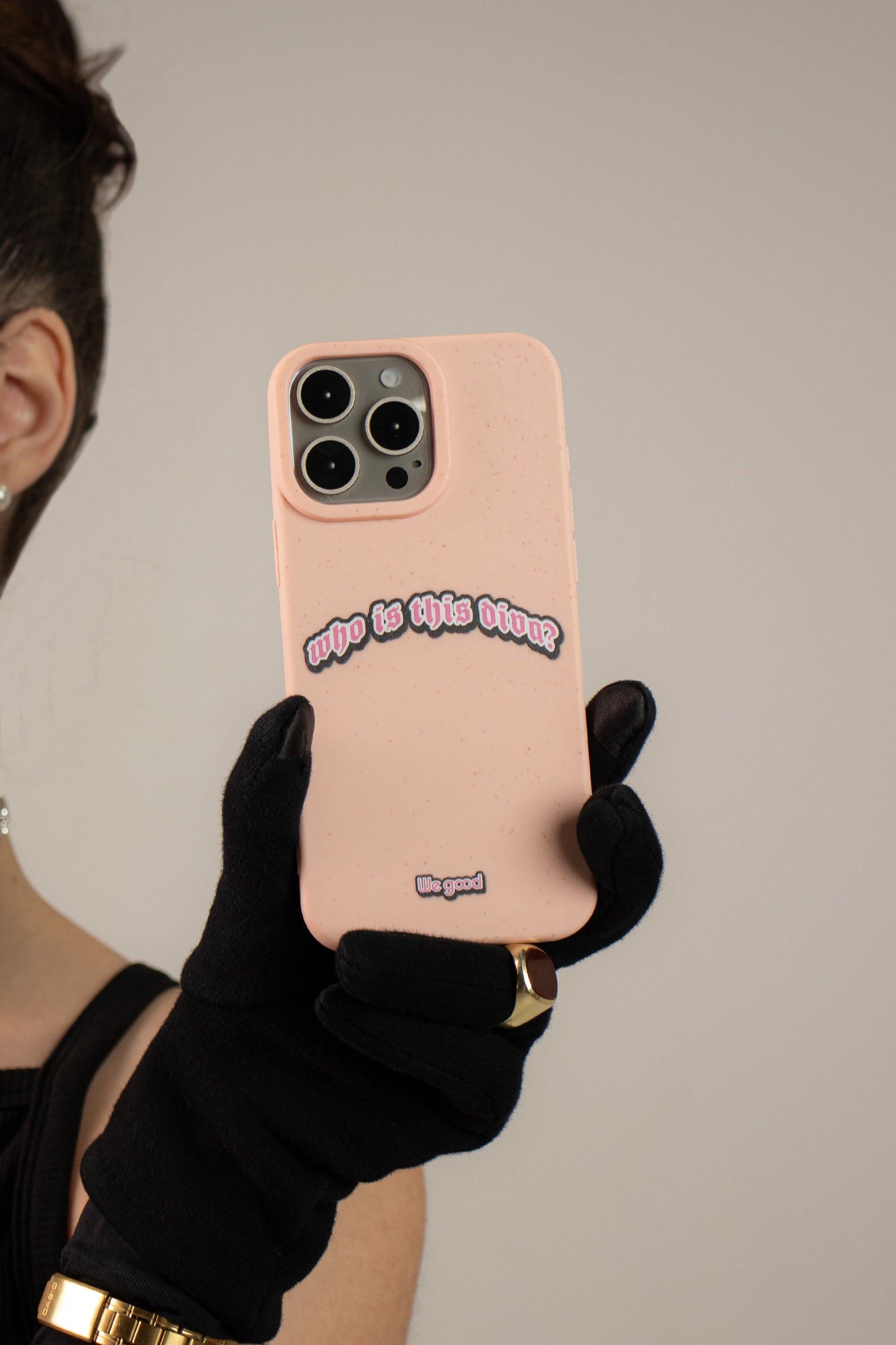 Woman With a black dress wearing black gloves holding a We Good Tpu iPhone case with wheat mix speckled in a flamingo pink color with a pink, white and black 'who is this diva?' gothic text print.