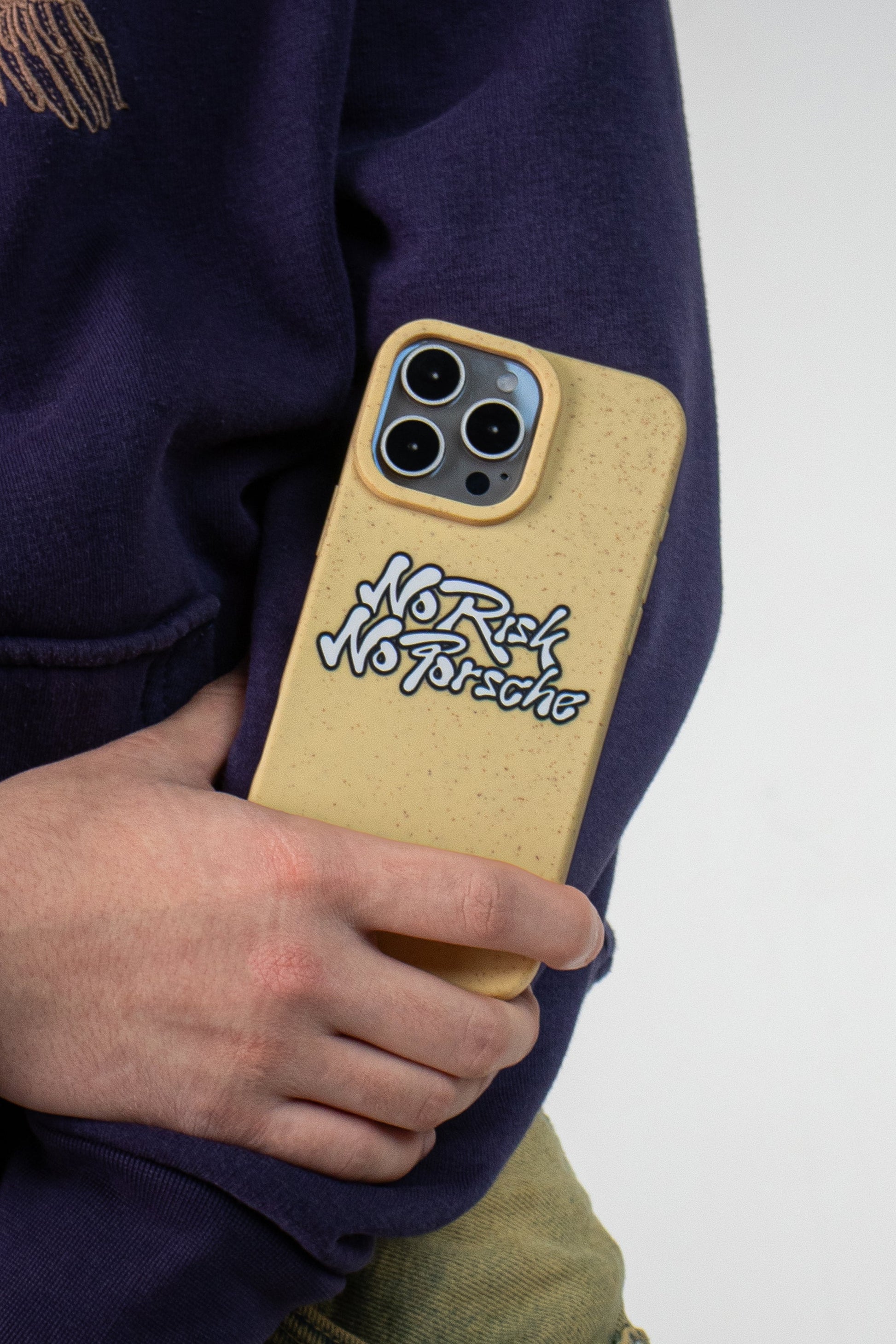 Man wearing a purple hoodie holding a We Good Tpu iPhone case with wheat mix speckled in a canary yellow color with black "No Risk No Porsche" bubble text print out of his pocket.