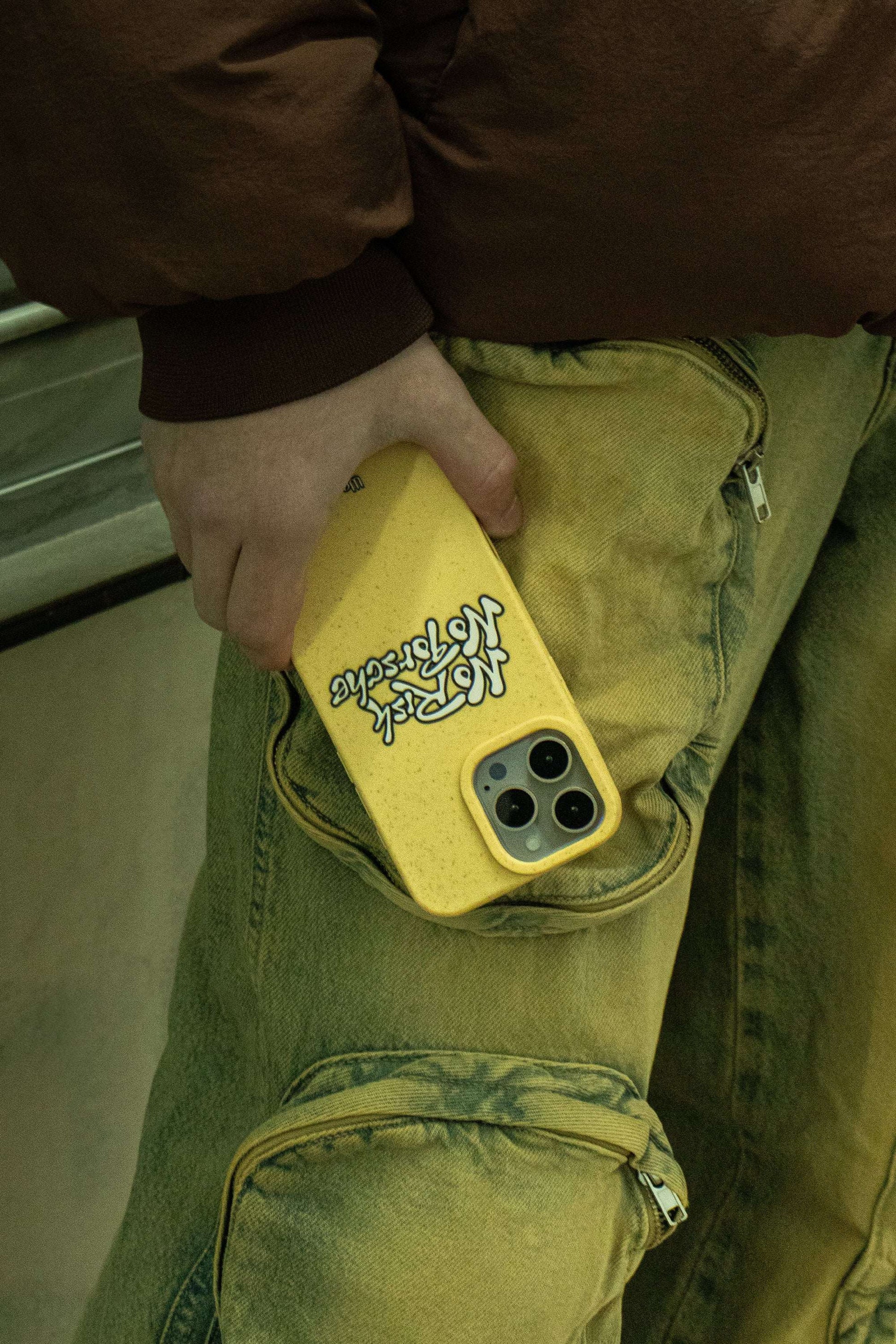 Man in a dimly lit alley wearing vintage jeans and brown jacket holding a We Good Tpu iPhone case with wheat mix speckled in a canary yellow color with black "No Risk No Porsche" bubble text print.