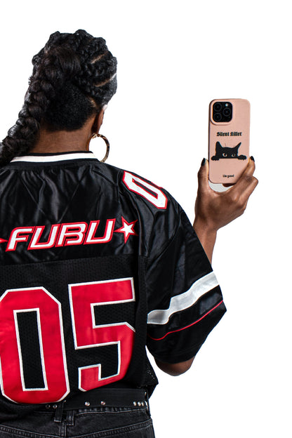 Woman with a black and red jersey holding a We Good Tpu iPhone case with wheat mix speckled in a flamingo pink color with a black 'silent killer' text in a gothic font and below it a black cats face creeping up and stalking. 