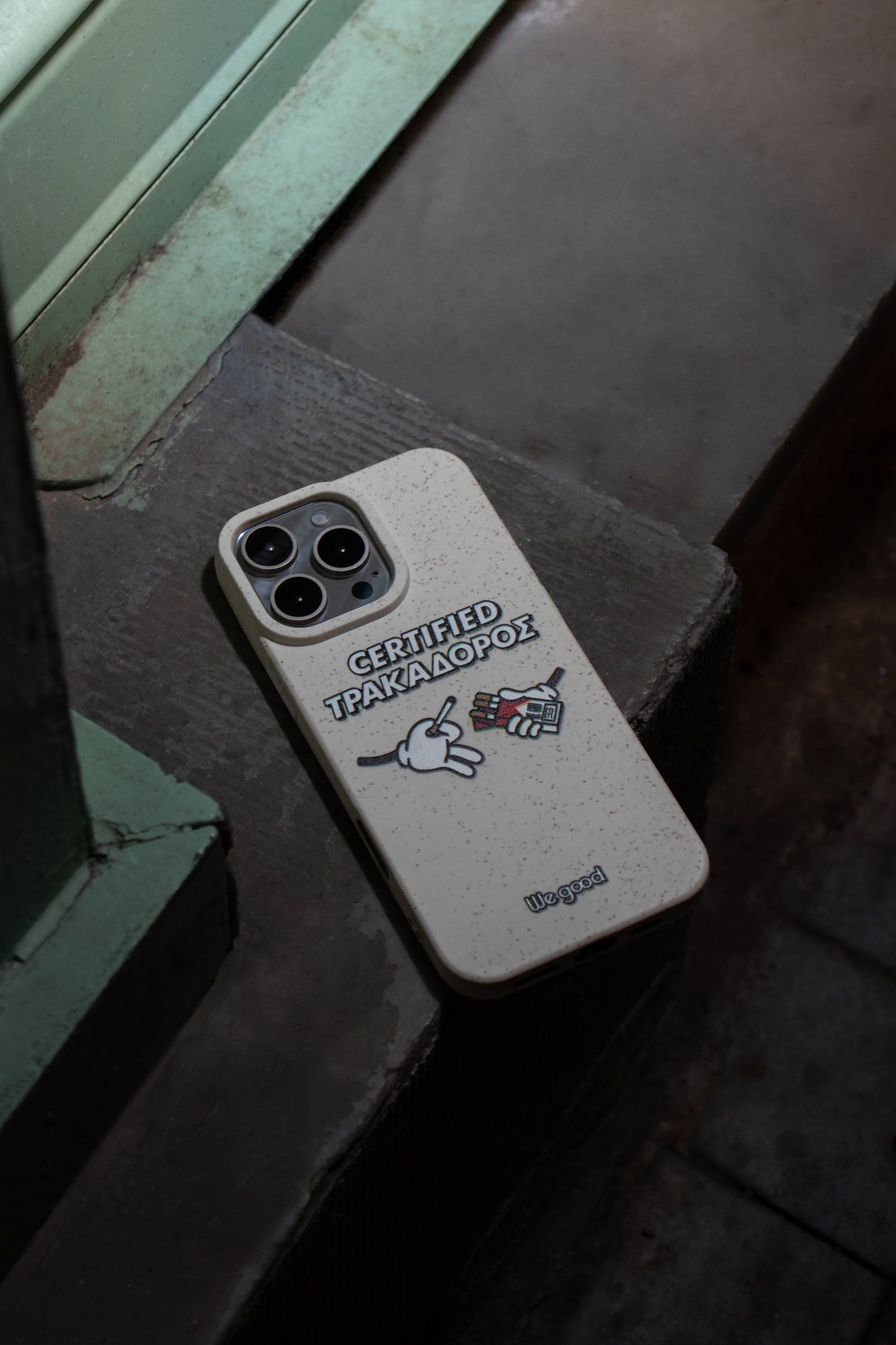 We Good Tpu iPhone case with wheat mix speckled in an off-white color with 'certified trakadoros' white and black bold text print and below it two cartoon hands with white gloves with one holding a red pack of cigarettes and the other hand taking one from the pack resting on the ground in a street setting at night.