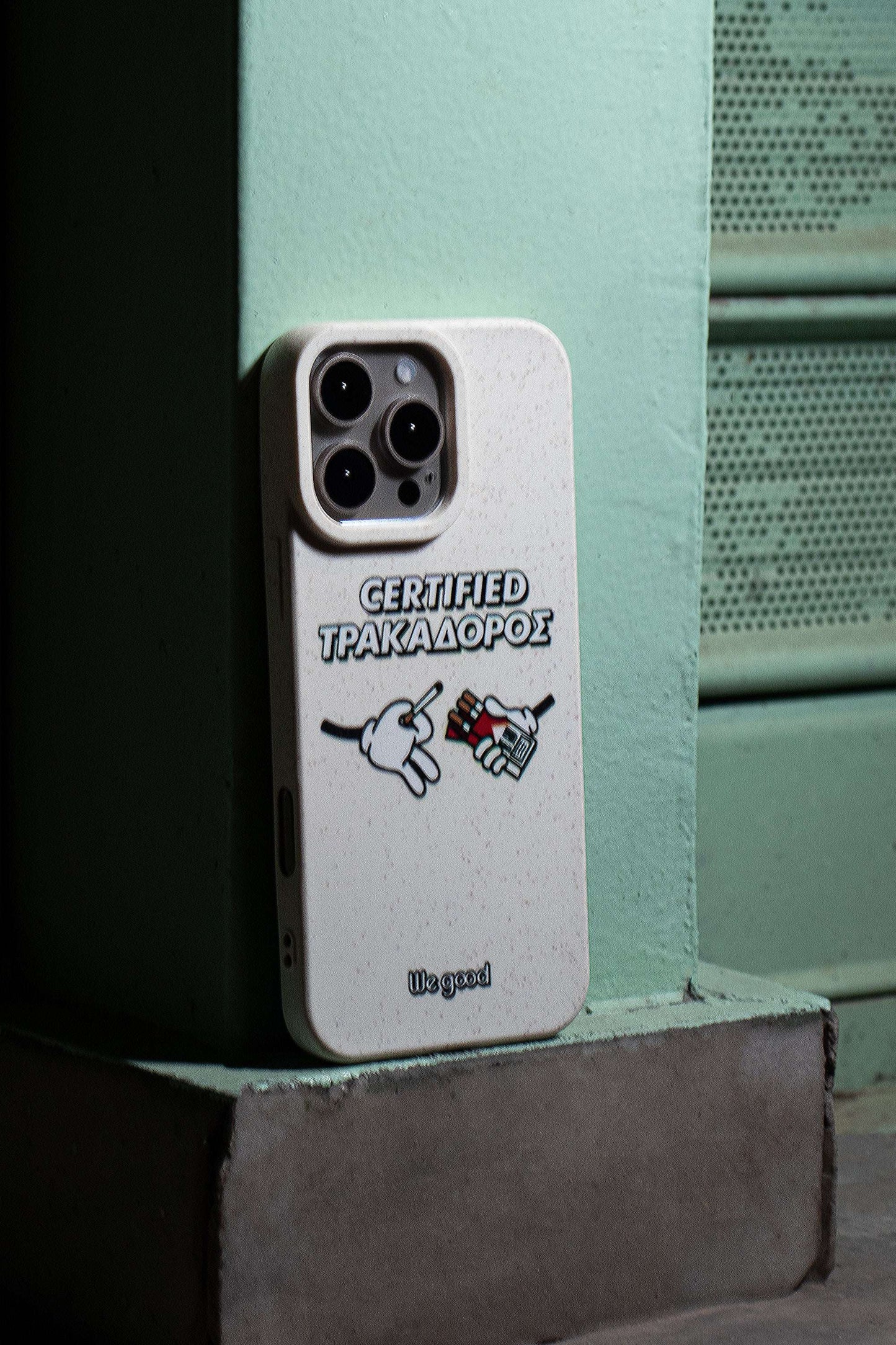 We Good Tpu iPhone case with wheat mix speckled in an off-white color with 'certified trakadoros' white and black bold text print and below it two cartoon hands with white gloves with one holding a red pack of cigarettes and the other hand taking one from the pack resting on the corner of a street at night.