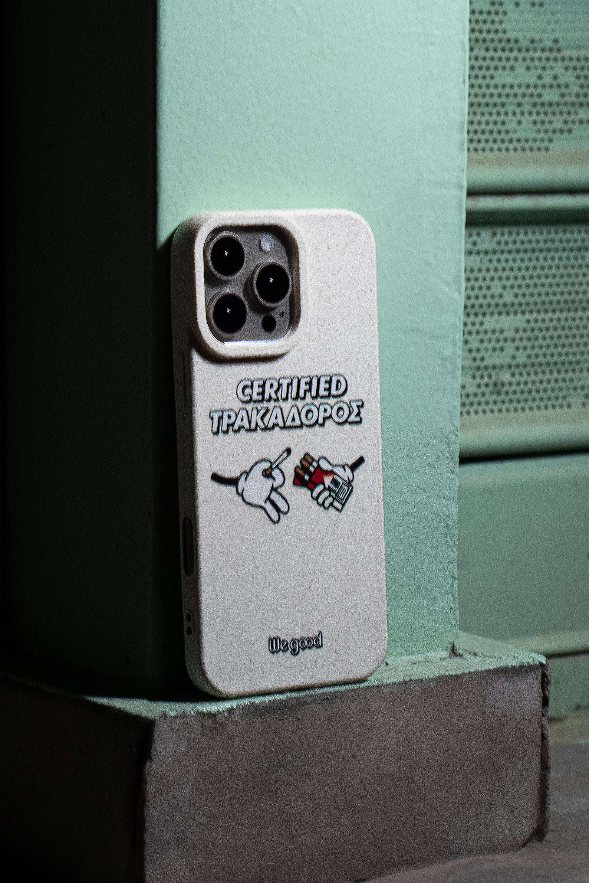 We Good Tpu iPhone case with wheat mix speckled in an off-white color with 'certified trakadoros' white and black bold text print and below it two cartoon hands with white gloves with one holding a red pack of cigarettes and the other hand taking one from the pack resting on the corner of a street at night.