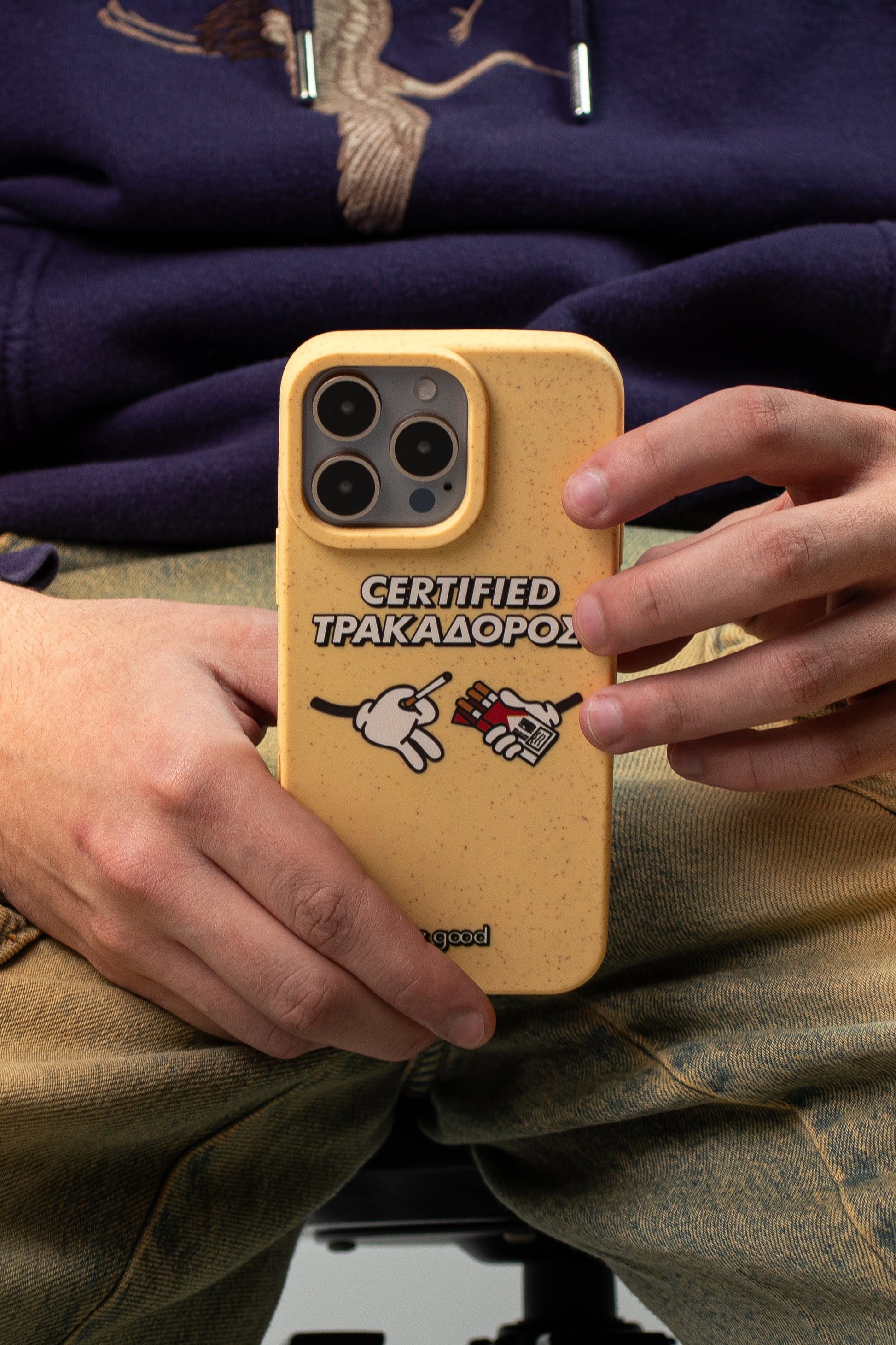 Man wearing vintage jeans and a purple hoodie holding a We Good Tpu iPhone case with wheat mix speckled in a canary yellow color with 'certified trakadoros' white and black bold text print and below it two cartoon hands with white gloves with one holding a red pack of cigarettes and the other hand taking one from the pack.