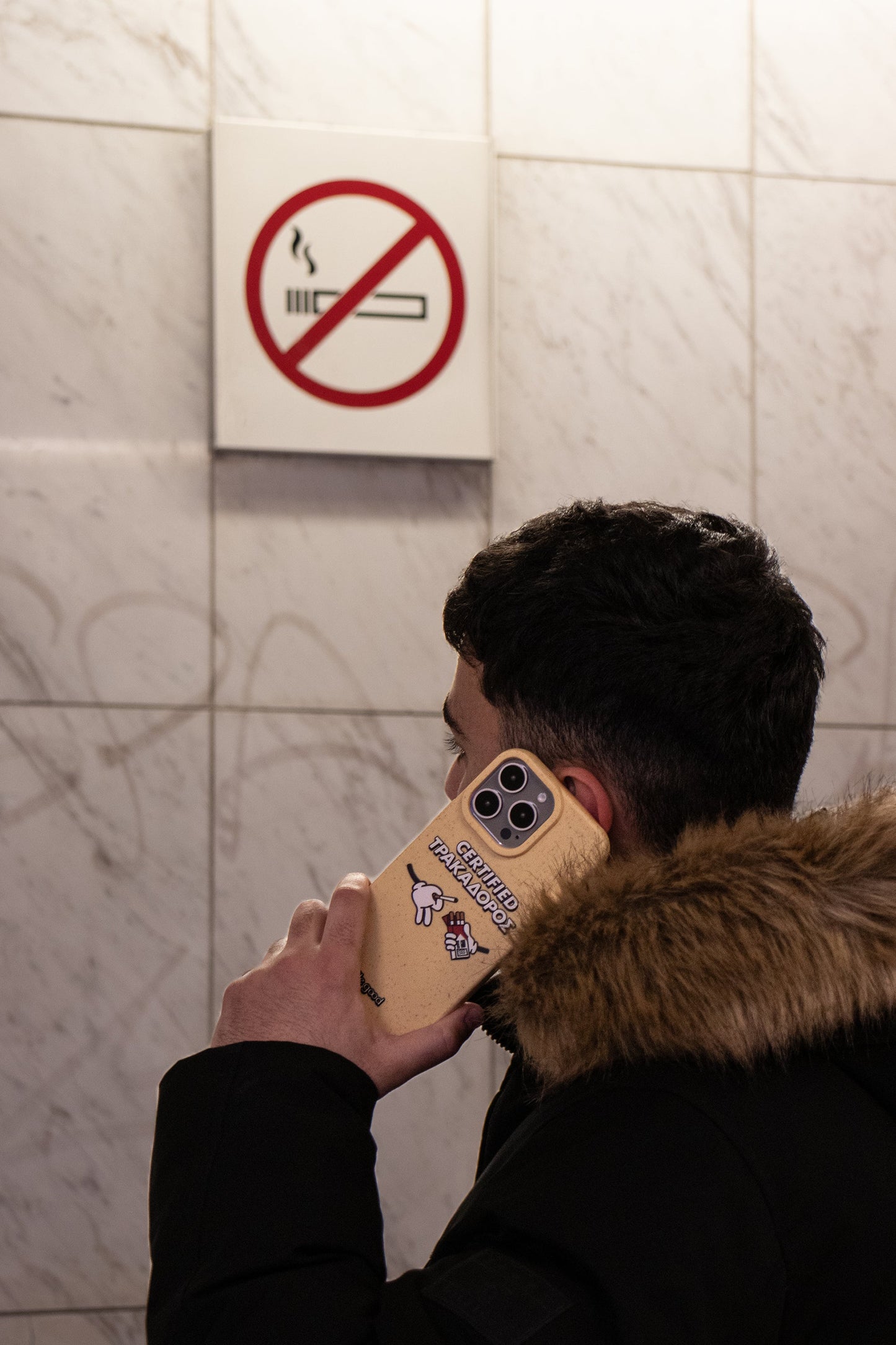 Man wearing a puffer jacket talking on the phone in a metro station below a no smoking sign, holding a We Good Tpu iPhone case with wheat mix speckled in a canary yellow color with 'certified trakadoros' white and black bold text print and below it two cartoon hands with white gloves with one holding a red pack of cigarettes and the other hand taking one from the pack. in his hand
