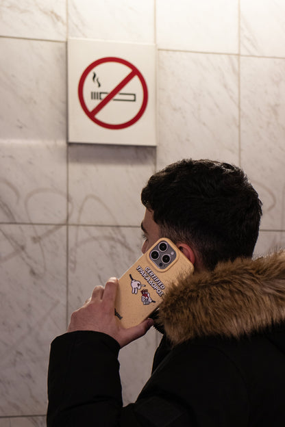 Man wearing a puffer jacket talking on the phone in a metro station below a no smoking sign, holding a We Good Tpu iPhone case with wheat mix speckled in a canary yellow color with 'certified trakadoros' white and black bold text print and below it two cartoon hands with white gloves with one holding a red pack of cigarettes and the other hand taking one from the pack. in his hand