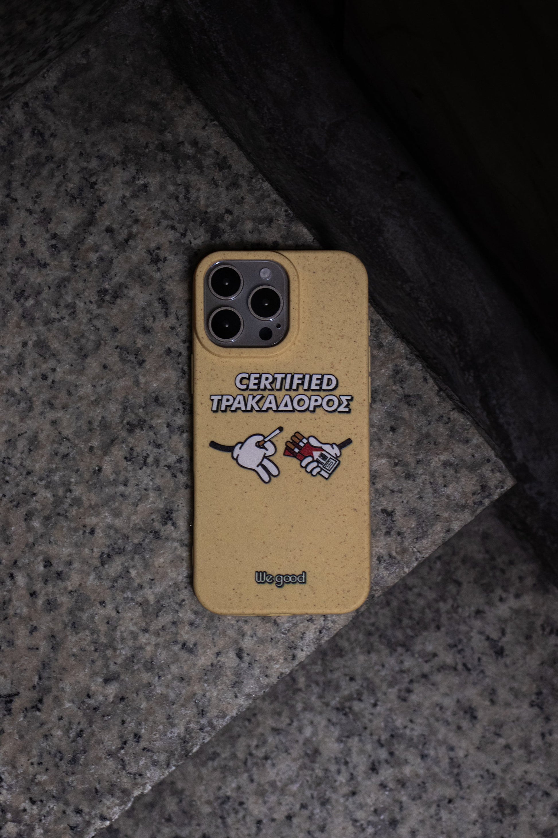 We Good Tpu iPhone case with wheat mix speckled in a canary yellow color with 'certified trakadoros' white and black bold text print and below it two cartoon hands with white gloves with one holding a red pack of cigarettes and the other hand taking one from the pack sitting on marble stairs at night from a top down view.