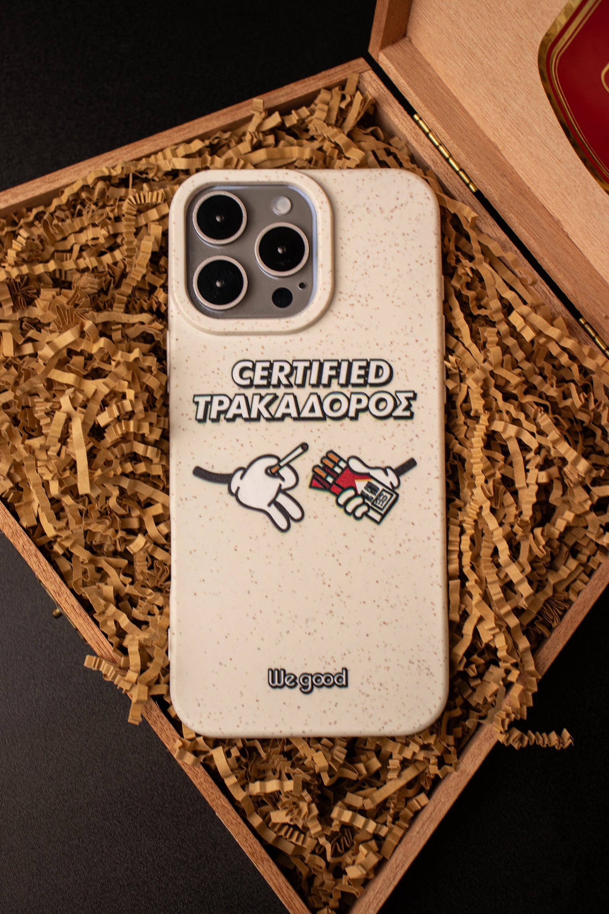 We Good Tpu iPhone case with wheat mix speckled in an off-white color with 'certified trakadoros' white and black bold text print and below it two cartoon hands with white gloves with one holding a red pack of cigarettes and the other hand taking one from the pack sitting inside a cigar case with brown paper below it to mimic tobacco leaves.